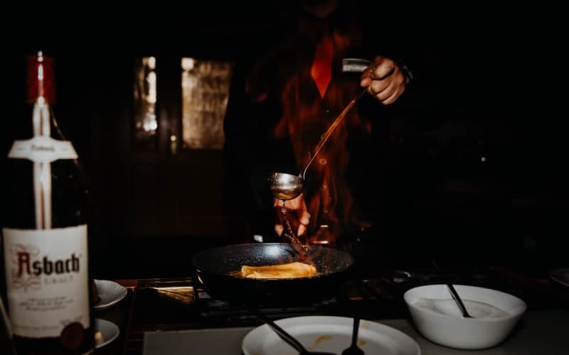 Chef flambéing crêpes in a pan with high flames, a bottle of Asbach in the background