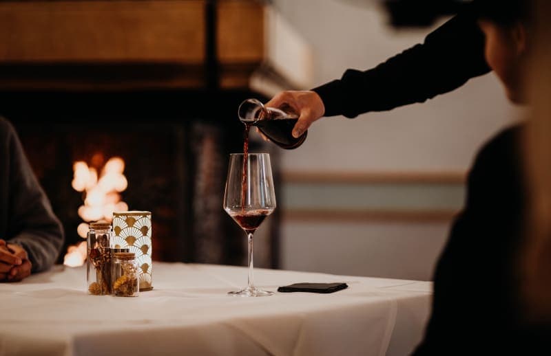 Un serveur verse du vin rouge dans un verre, à une table élégamment dressée devant une cheminée allumée
